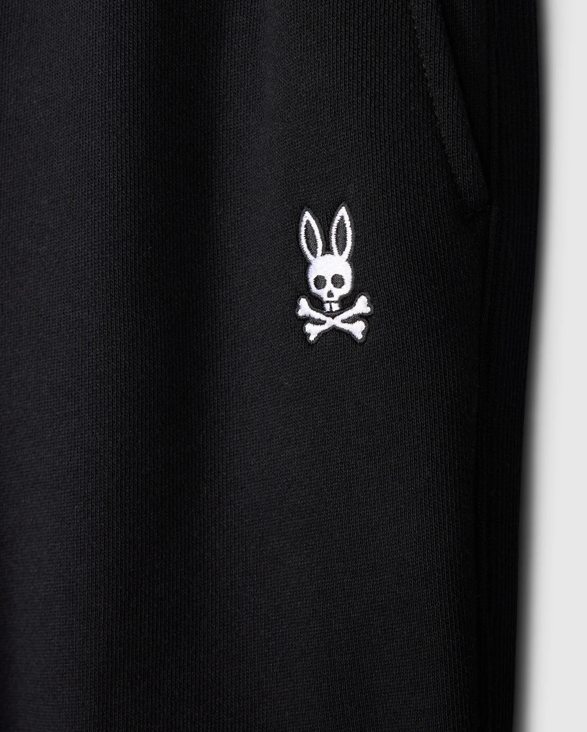 Close-up of the black fabric on Psycho Bunny's KIDS LEVY ESSENTIAL FLEECE SWEATPANT (B0P721D200), highlighting a white embroidered bunny skull and crossbones logo. The cozy fleece features a textured, ribbed look with a visible seam for added style.