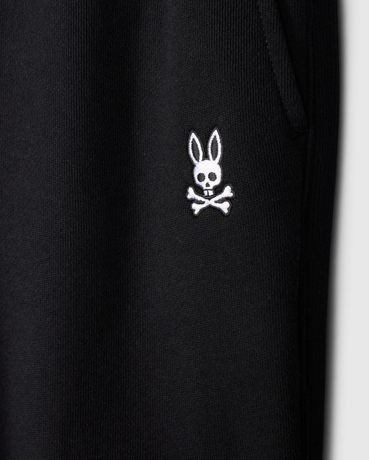 Close-up of the black fabric on Psycho Bunny's KIDS LEVY ESSENTIAL FLEECE SWEATPANT (B0P721D200), highlighting a white embroidered bunny skull and crossbones logo. The cozy fleece features a textured, ribbed look with a visible seam for added style.