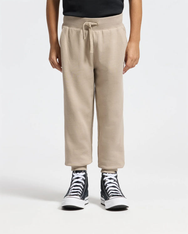 A person in Psycho Bunny's Kids Levy Essential Fleece Sweatpants stands against a plain white background. The beige sweatpants, featuring a drawstring waist, are paired with black high-top sneakers and a sleek black top, effortlessly blending comfort with style.