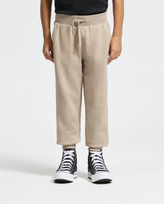 A person in Psycho Bunny's Kids Levy Essential Fleece Sweatpants stands against a plain white background. The beige sweatpants, featuring a drawstring waist, are paired with black high-top sneakers and a sleek black top, effortlessly blending comfort with style.