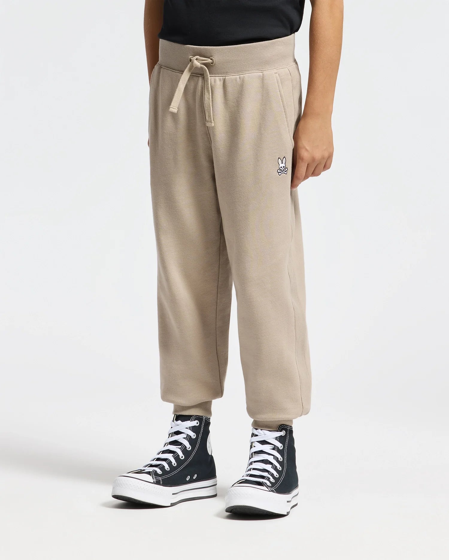 A person is wearing Psycho Bunny's KIDS LEVY ESSENTIAL FLEECE SWEATPANT with a drawstring and small leg design, paired with black high-top sneakers and a black top, all set against a white background.