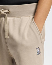 A person wearing Psycho Bunny's KIDS LEVY ESSENTIAL FLEECE SWEATPANT featuring a white bunny and crossbones logo on the pocket, with their hand in the pocket and a black shirt.