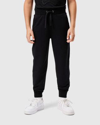 A person standing against a plain background, wearing Psycho Bunny's KIDS WALTER LIGHTWEIGHT SWEATPANT (B0P860D200) made from breathable cotton-blend with a drawstring waist and white sneakers. The sweatpants have elastic cuffs at the ankles, perfect for casual wear. The upper portion of the person's outfit is not fully visible.