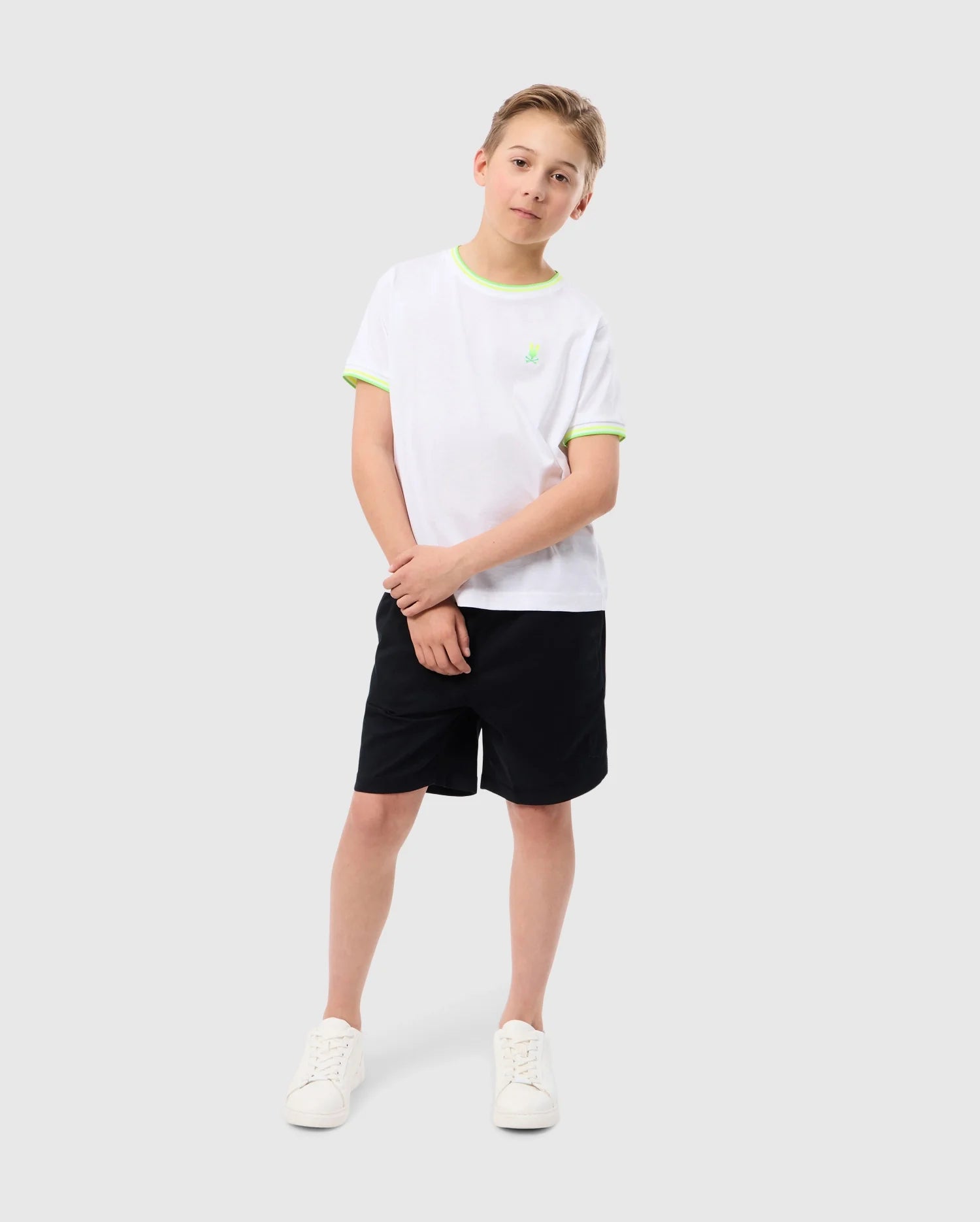 A young boy stands against a plain background, wearing a slim-fit white tee with green trim and an embroidered Psycho Bunny logo, paired with Psycho Bunny's KIDS WILLIS STRETCH TENCEL SHORT in black and white sneakers. He smiles slightly, hands together in front.