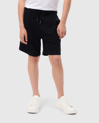 A person is seen against a light gray backdrop, wearing the Psycho Bunny KIDS WILLIS STRETCH TENCEL SHORT (B0R239Y1WB) in black, featuring a slim-fit design with an embroidered bunny logo and drawstring waist, paired with white sneakers. The upper body isn't visible.