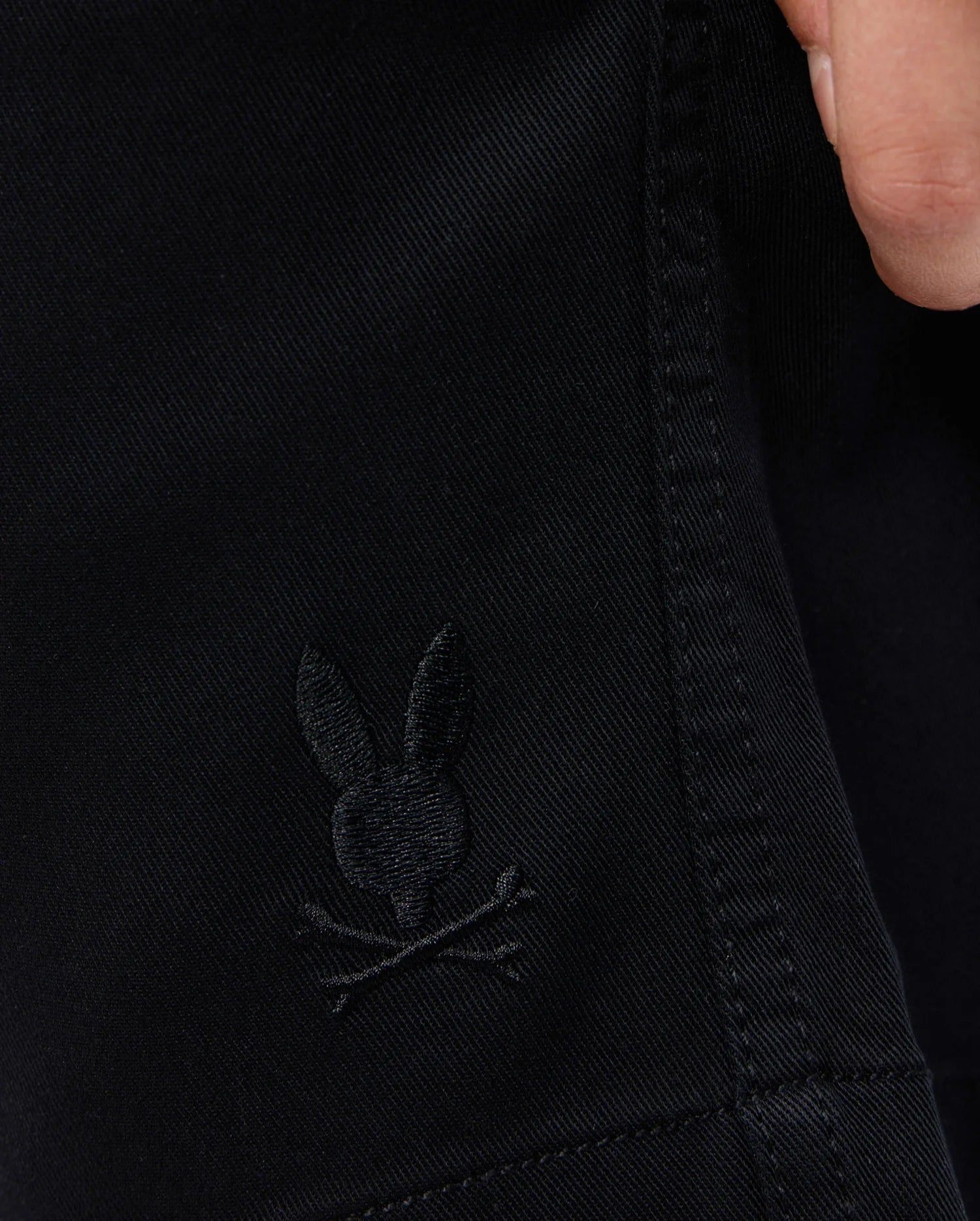 Close-up of black Psycho Bunny KIDS WILLIS STRETCH TENCEL SHORT (B0R239Y1WB) fabric, featuring an embroidered bunny head with crossed bones. A hand is partially visible at the top right corner, enhancing the intricate design.