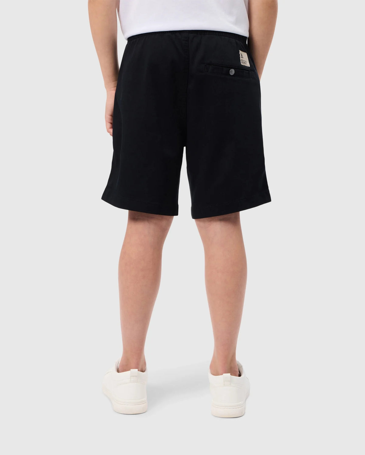 A person in Psycho Bunny's black KIDS WILLIS STRETCH TENCEL SHORT - B0R239Y1WB and white sneakers stands with their back to the camera. Their white shirt enhances the minimalist style, and an embroidered bunny logo decorates a buttoned back pocket. The background is plain white.