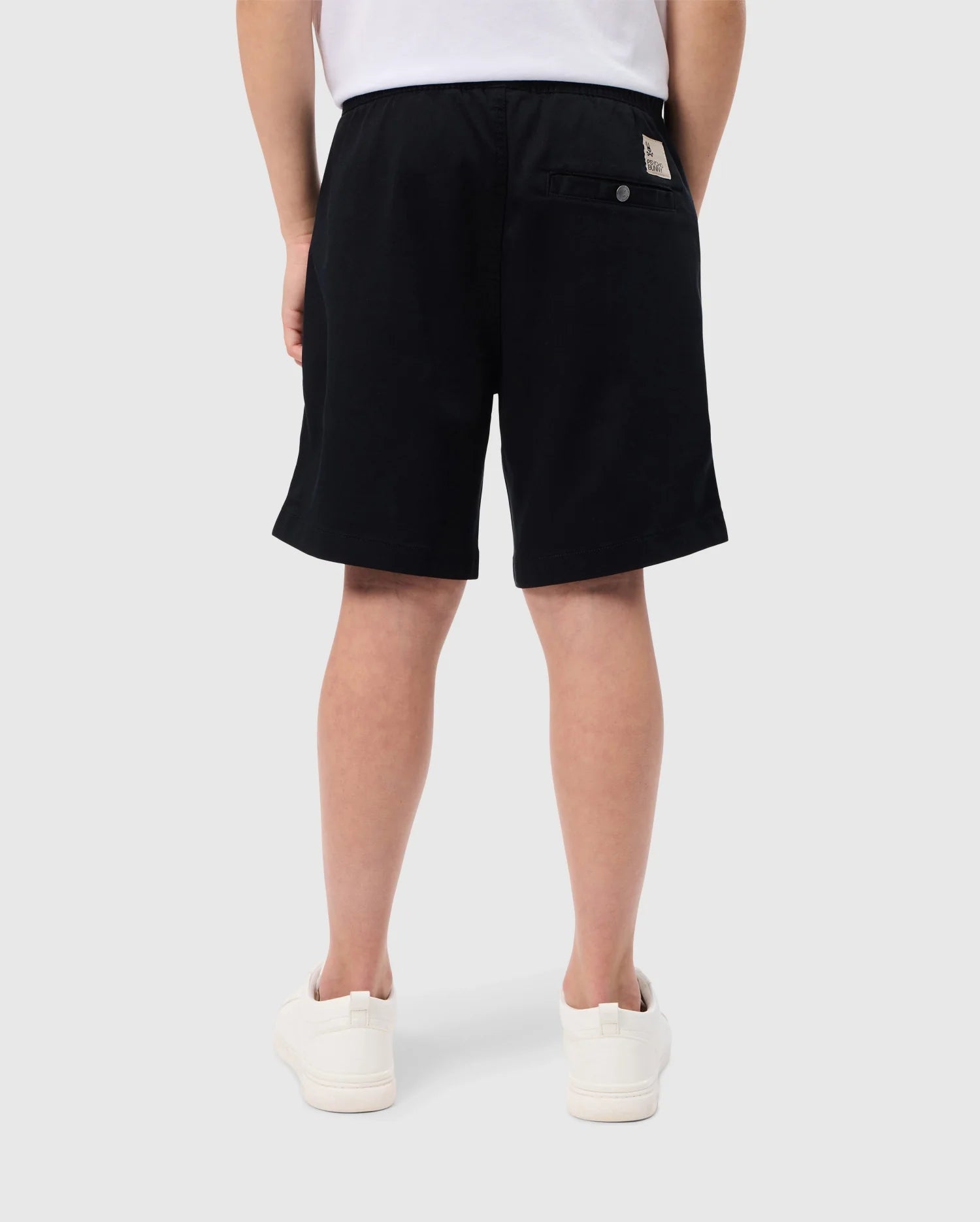 A person in Psycho Bunny's black KIDS WILLIS STRETCH TENCEL SHORT - B0R239Y1WB and white sneakers stands with their back to the camera. Their white shirt enhances the minimalist style, and an embroidered bunny logo decorates a buttoned back pocket. The background is plain white.