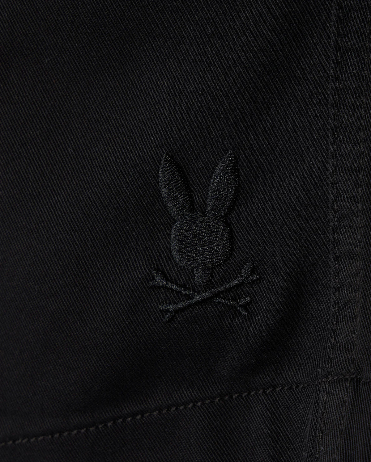 Close-up of the KIDS WILLIS STRETCH TENCEL SHORT by Psycho Bunny, featuring black fabric with an embroidered Bunny Logo—a stylized rabbit head and ears above crossed bones in a playful skull and crossbones motif. The Slim Fit design includes visible stitching for a sleek look.