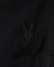 Close-up of the KIDS WILLIS STRETCH TENCEL SHORT by Psycho Bunny, featuring black fabric with an embroidered Bunny Logo—a stylized rabbit head and ears above crossed bones in a playful skull and crossbones motif. The Slim Fit design includes visible stitching for a sleek look.