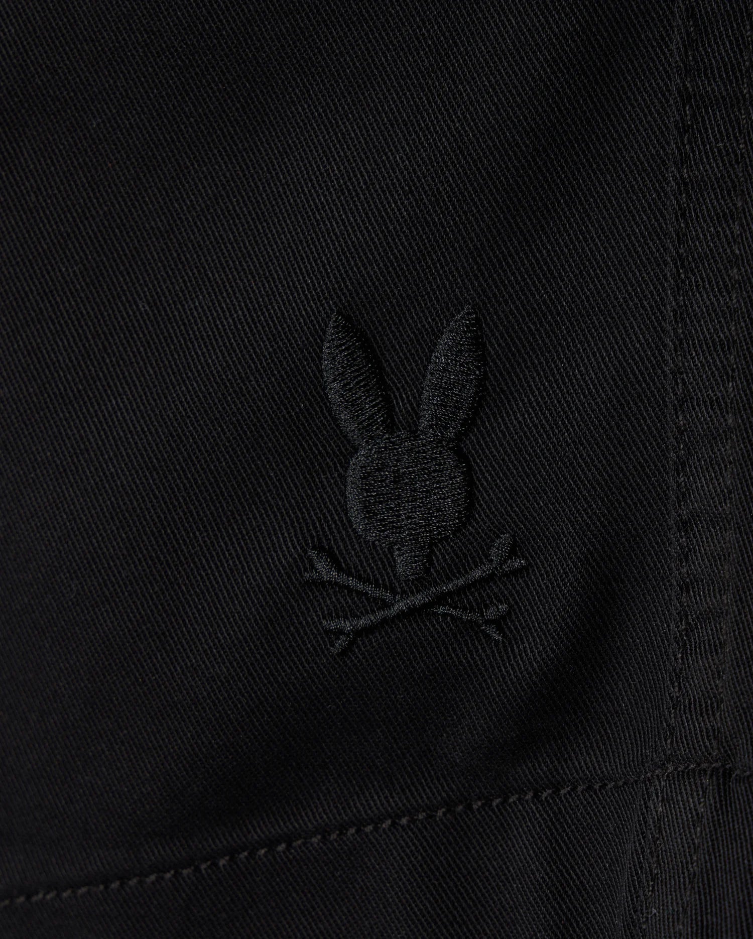 Close-up of the KIDS WILLIS STRETCH TENCEL SHORT by Psycho Bunny, featuring black fabric with an embroidered Bunny Logo—a stylized rabbit head and ears above crossed bones in a playful skull and crossbones motif. The Slim Fit design includes visible stitching for a sleek look.