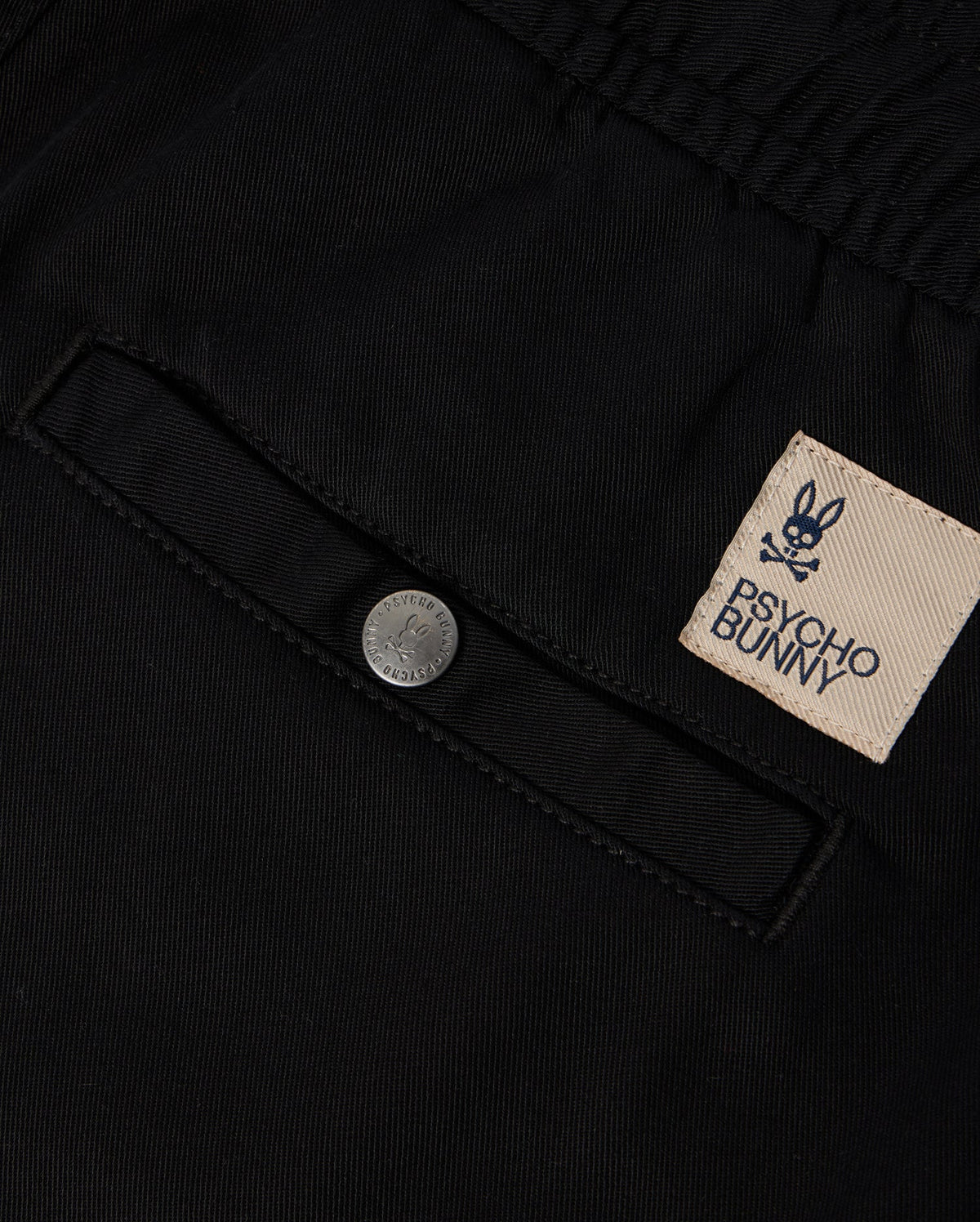 Close-up of a black fabric with a buttoned pocket, featuring an embroidered Psycho Bunny label with the bunny skull logo. The KIDS WILLIS STRETCH TENCEL SHORT (B0R239Y1WB) offers a slim fit design that incorporates Stretch TENCEL™ for comfort and style.