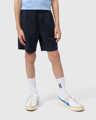 A person in a light blue shirt stands wearing Psycho Bunny's Kids' Willis Stretch Tencel Short (B0R239Y1WB) in dark blue, paired with white socks featuring a small design. Their shoes are white with blue stripes and beige soles, set against a plain light gray background.
