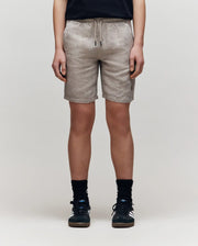 A person stands against a plain light background, shown from the waist down, wearing Psycho Bunny KIDS linen-cotton shorts with a drawstring waist, paired with a black shirt, black socks, and black sneakers.