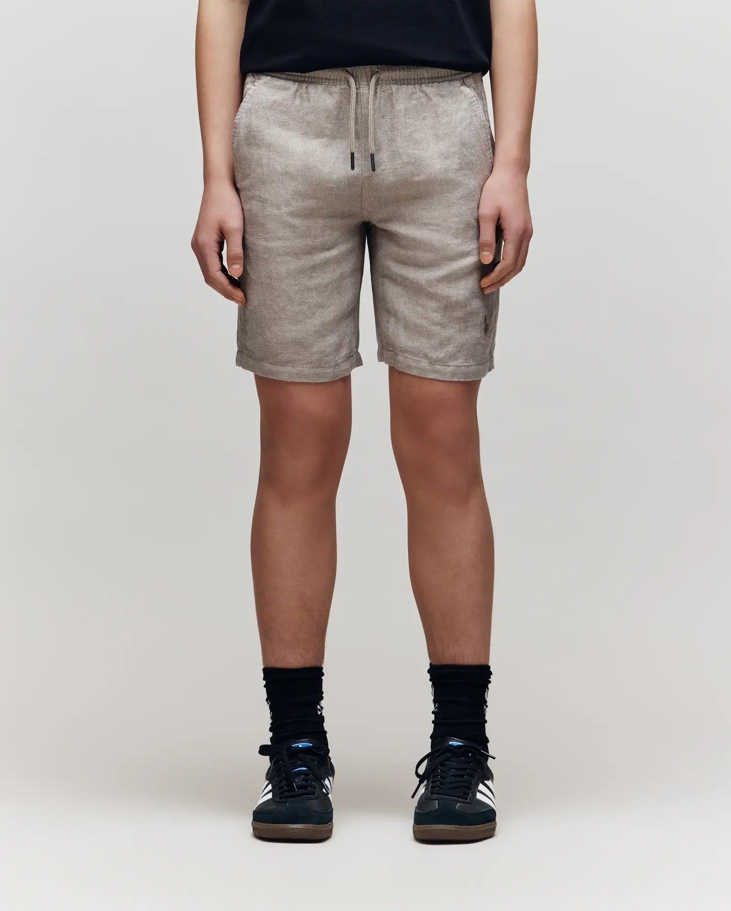 A person stands against a plain light background, shown from the waist down, wearing Psycho Bunny KIDS linen-cotton shorts with a drawstring waist, paired with a black shirt, black socks, and black sneakers.