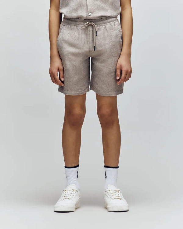 A person, visible from chest down, wears Psycho Bunny KIDS beige drawstring shorts and a matching short-sleeve shirt with white crew socks and sneakers, standing against a light gray background.