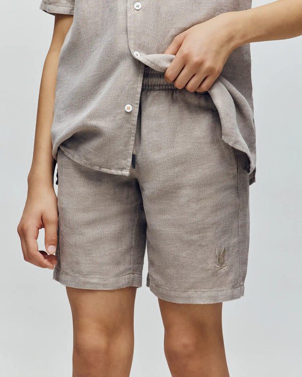 A person wears Psycho Bunny's KIDS beige short-sleeve button-up shirt with matching breezy shorts, standing against a plain background and lifting the shirt to show the shorts' elastic waistband.