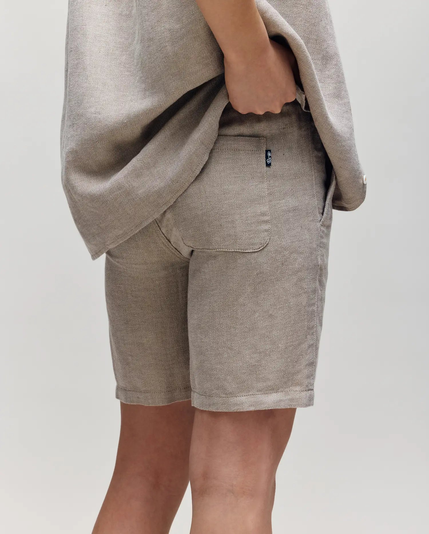 A person wears Psycho Bunny KIDS beige breezy shorts with a matching shirt, standing against a plain light background with one hand in their back pocket.