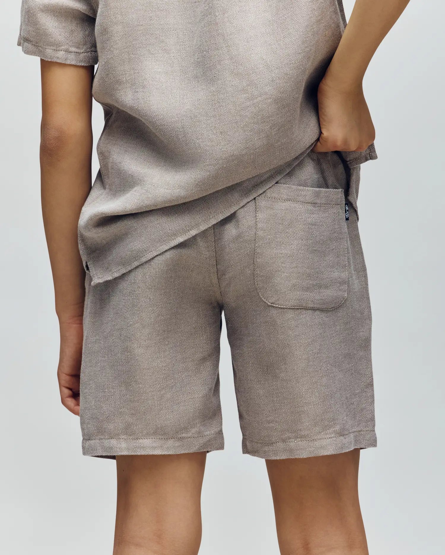 A person faces away from the camera, wearing Psycho Bunny's KIDS light beige textured short-sleeve shirt and matching linen-cotton drawstring shorts. One hand is in the back pocket against a plain light gray background.
