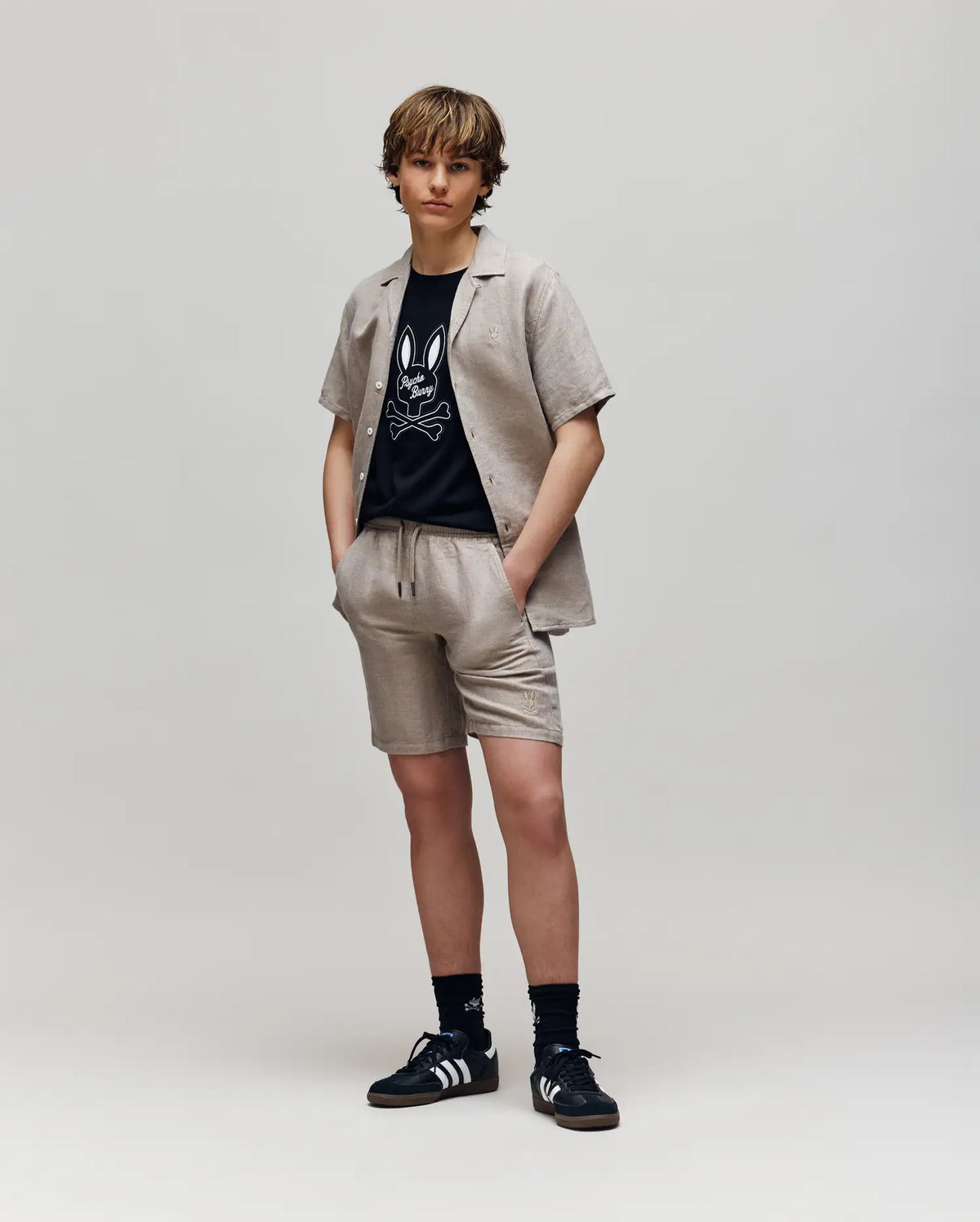 A young person stands against a plain background wearing Psycho Bunny KIDS beige linen-cotton drawstring shorts and matching short-sleeve shirt over a black cartoon graphic tee, paired with black Adidas sneakers and socks.