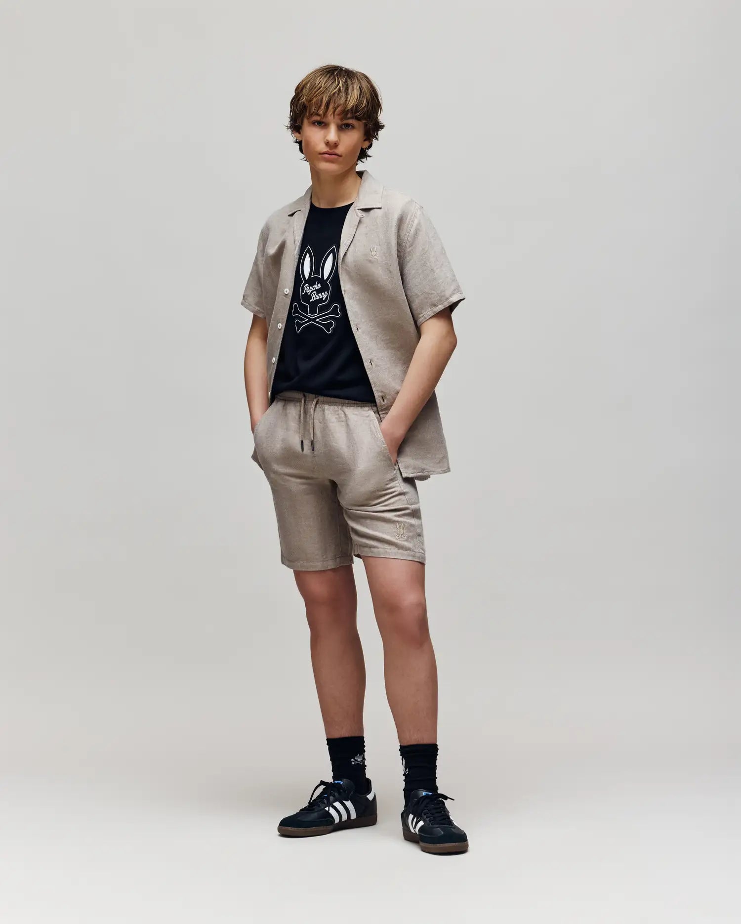 A young person stands against a plain background wearing Psycho Bunny KIDS beige linen-cotton drawstring shorts and matching short-sleeve shirt over a black cartoon graphic tee, paired with black Adidas sneakers and socks.