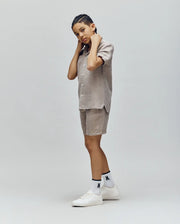 A person with short braids poses in Psycho Bunny KIDS beige short-sleeve shirt and linen-cotton drawstring shorts, white graphic socks, and white sneakers against a plain light gray background.
