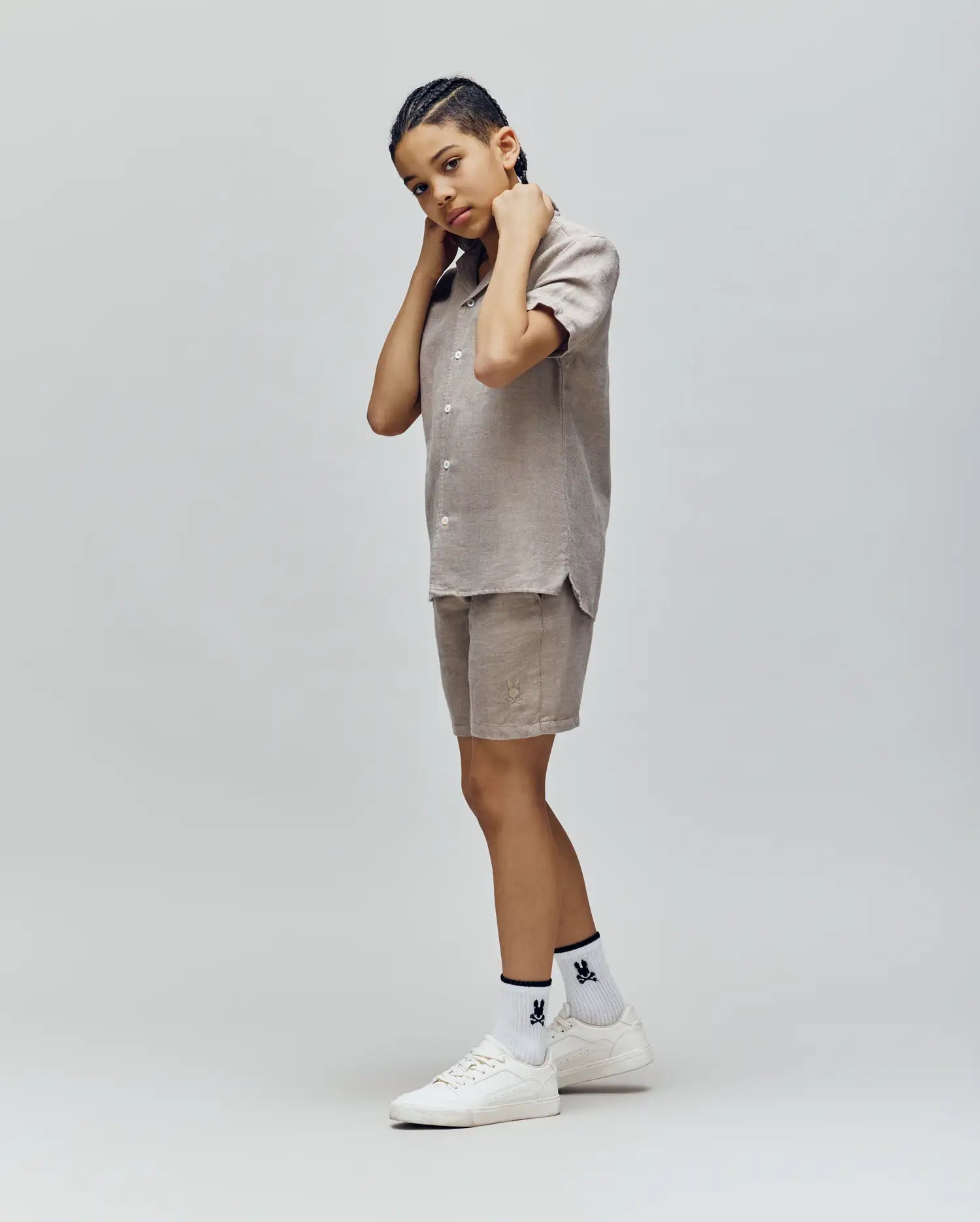 A person with short braids poses in Psycho Bunny KIDS beige short-sleeve shirt and linen-cotton drawstring shorts, white graphic socks, and white sneakers against a plain light gray background.