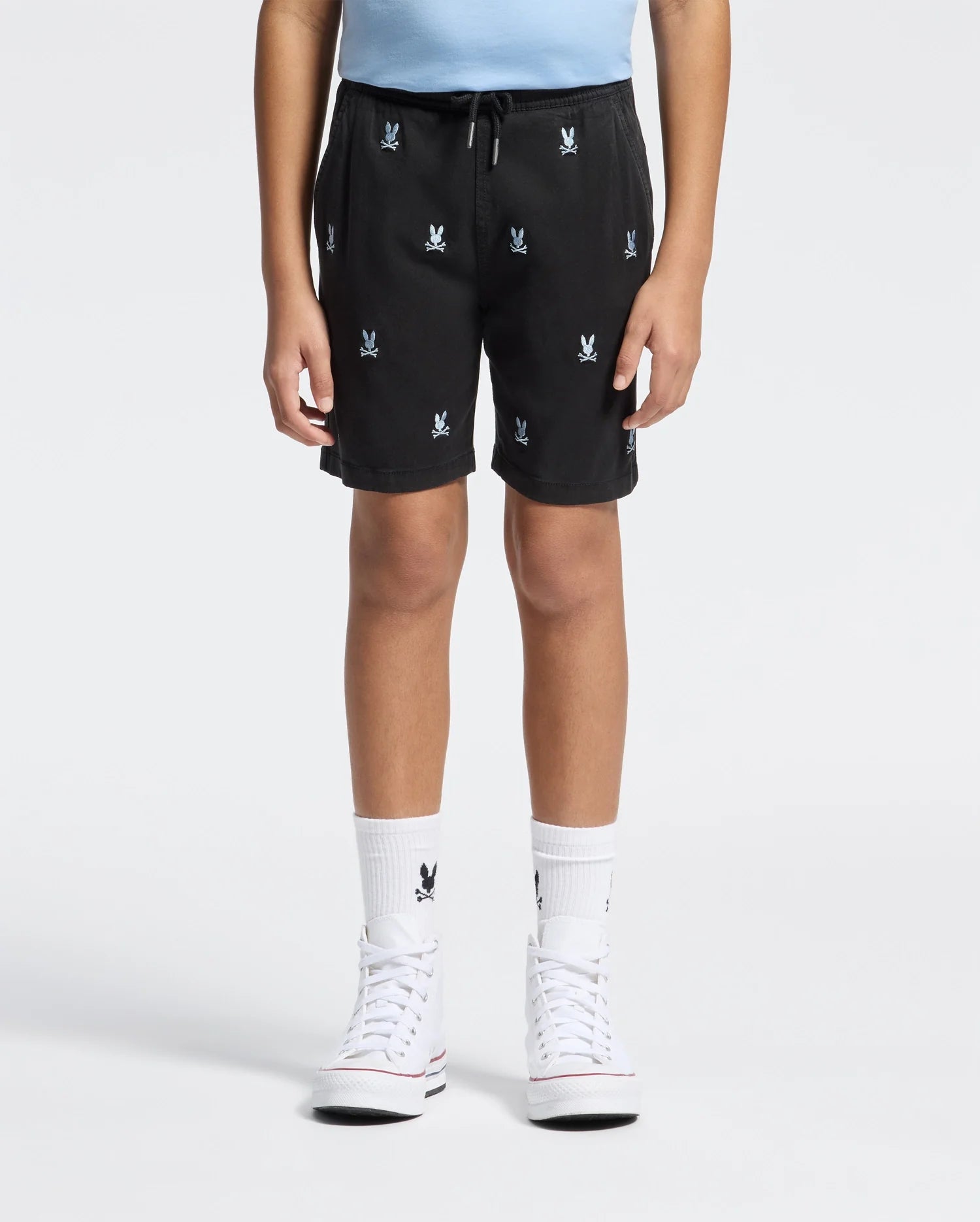 A child wears Psycho Bunny KIDS ALL OVER WILLIS STRETCH TENCEL SHORTS (B0R559F200) featuring white bunny logos, a light blue shirt, matching bunny socks, and white high-top sneakers, standing against a plain white background.