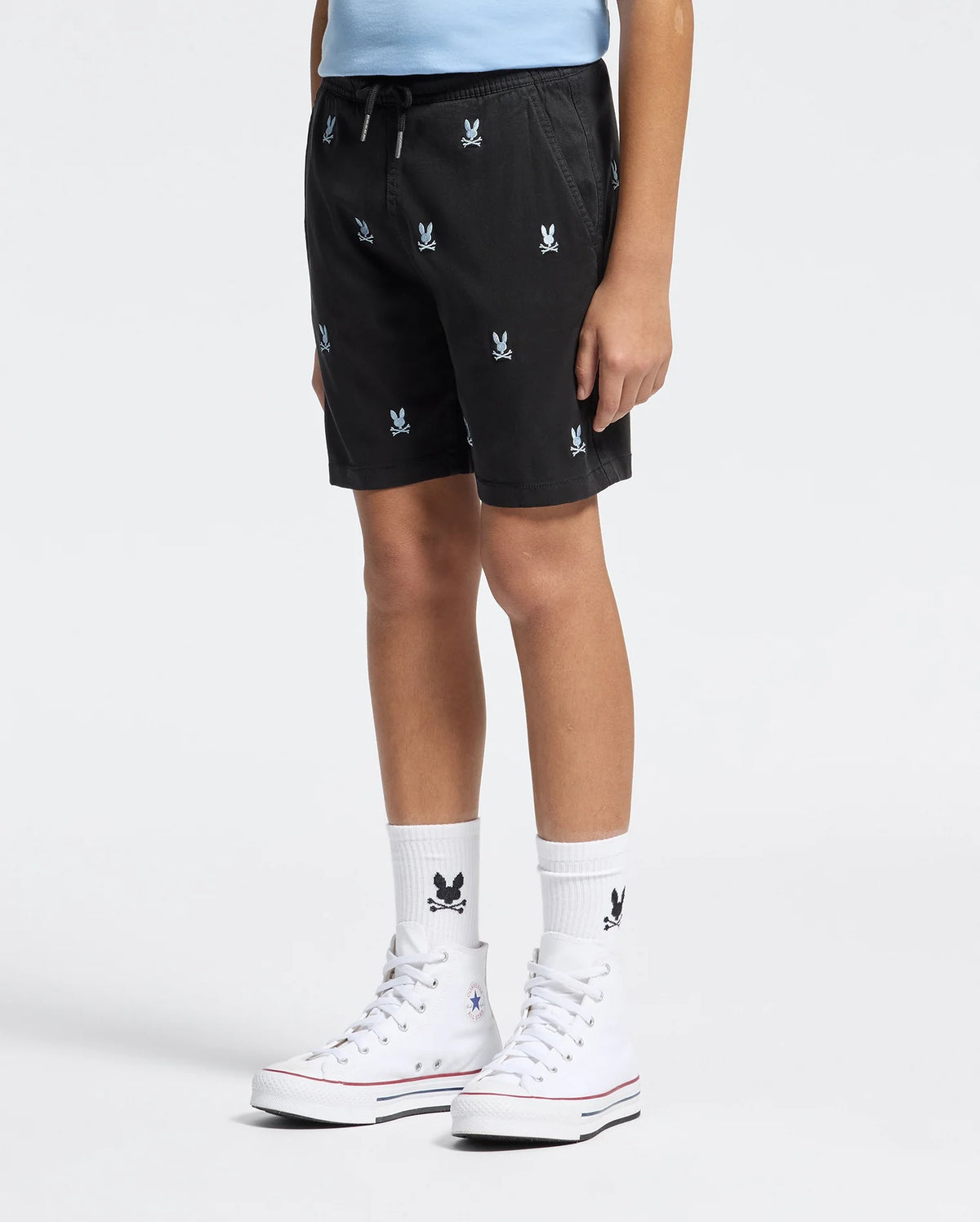 A person wears Psycho Bunny KIDS ALL OVER WILLIS STRETCH TENCEL SHORT - B0R559F200 in black with an elasticized waist and white embroidered logos, paired with a light blue shirt, white socks, and white high-top sneakers against a plain background.