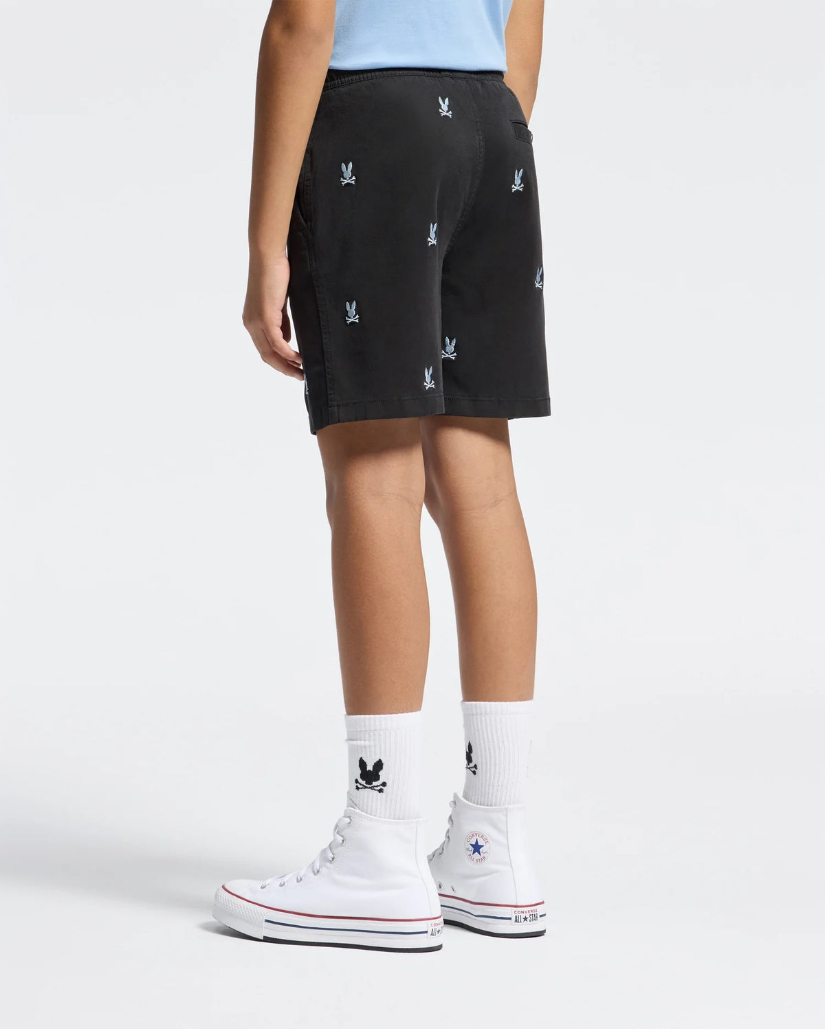 A person wears Psycho Bunny KIDS ALL OVER WILLIS STRETCH TENCEL SHORTS (B0R559F200) with a bunny pattern and elastic waist, styled with white crew socks featuring a black bunny logo and white high-top Converse sneakers against a white background.