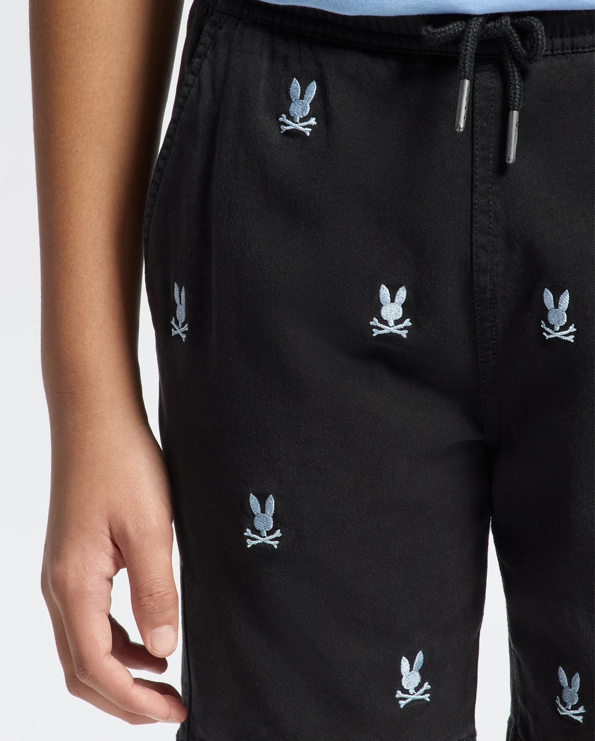 A person wears Psycho Bunny KIDS ALL OVER WILLIS STRETCH TENCEL SHORTS (B0R559F200) with a bunny and crossbones embroidery, elasticized drawstring waist. Only the lower torso and one hand are visible.