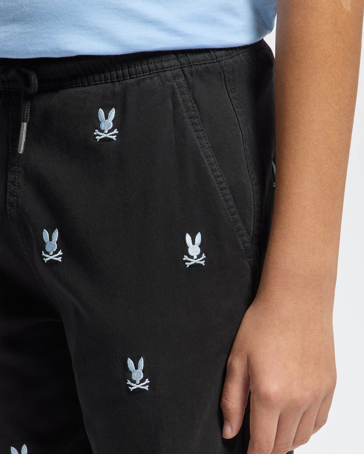 Close-up of a child wearing Psycho Bunny KIDS ALL OVER WILLIS STRETCH TENCEL SHORTS (B0R559F200) in black with an elastic waist and small white embroidered bunny head and crossbones, standing with one hand at their side; torso and arm partially visible.