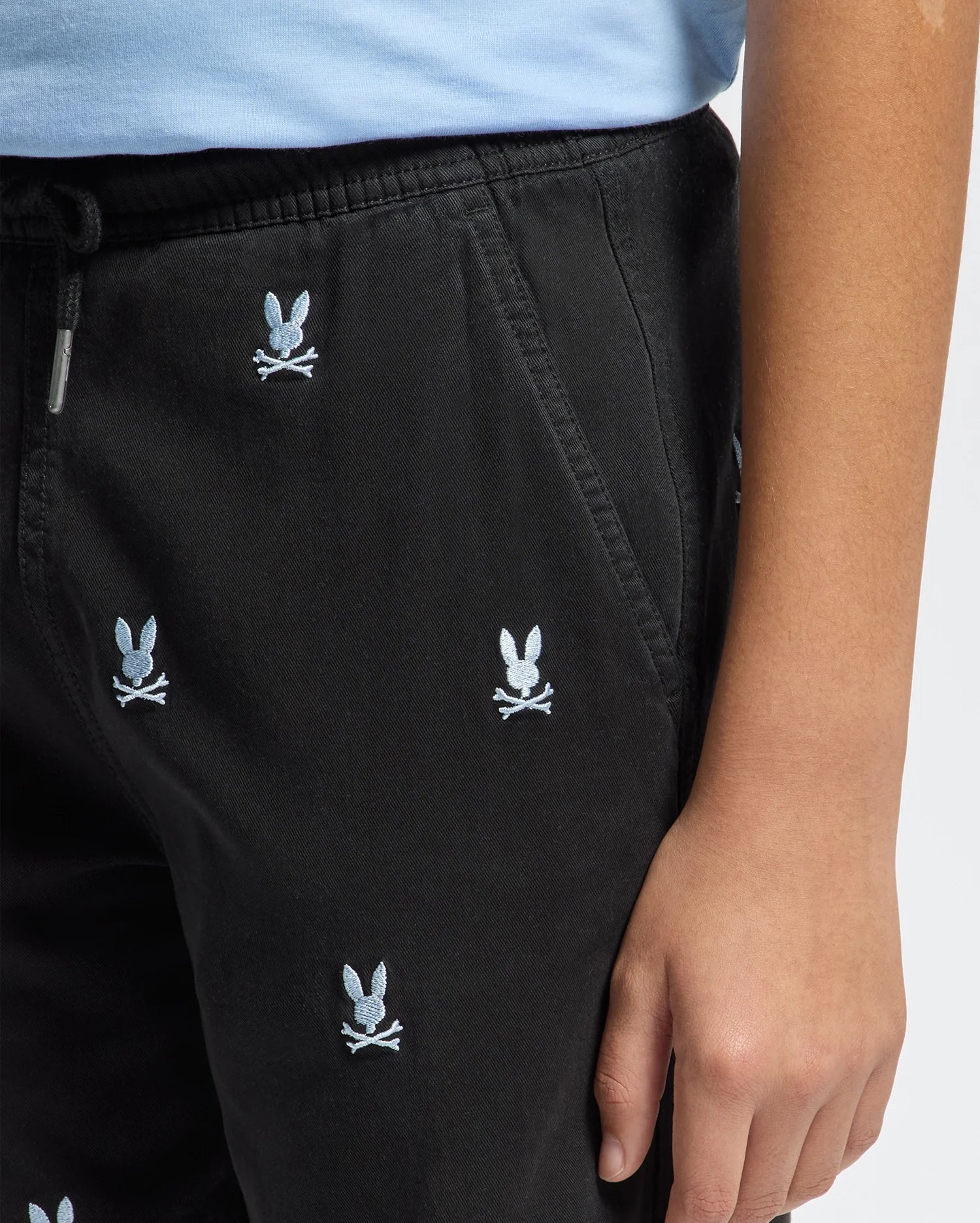 Close-up of a child wearing Psycho Bunny KIDS ALL OVER WILLIS STRETCH TENCEL SHORTS (B0R559F200) in black with an elastic waist and small white embroidered bunny head and crossbones, standing with one hand at their side; torso and arm partially visible.