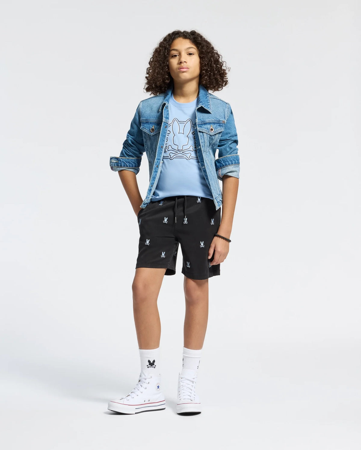 A young person with curly hair wears a Psycho Bunny KIDS ALL OVER WILLIS STRETCH TENCEL SHORT (B0R559F200), paired with a denim jacket, light blue graphic tee, white socks, and white high-top sneakers against a plain white background.
