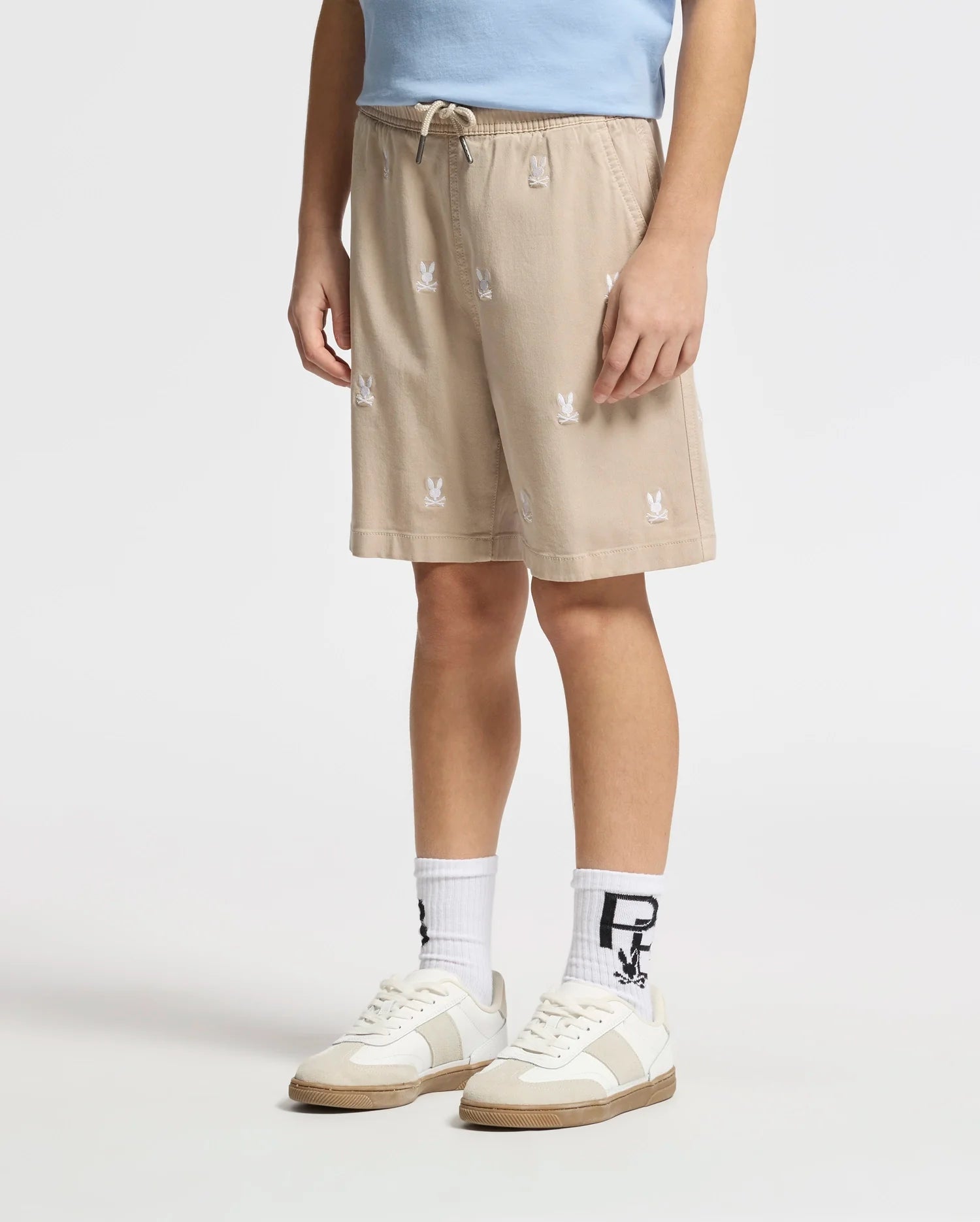 Wearing Psycho Bunny's KIDS ALL OVER WILLIS STRETCH TENCEL SHORT in regular fit tan with subtle white logos, paired with a light blue shirt, white socks with black lettering, and beige-accented white sneakers sets an effortlessly stylish tone against a plain white background.