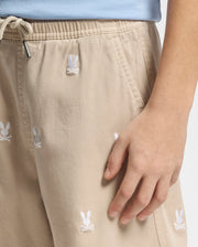 A person sports the Psycho Bunny KIDS ALL OVER WILLIS STRETCH TENCEL SHORT - B0R559F200 in beige, adorned with white bunny motifs, styled casually with a light blue top. The regular fit shorts provide comfort as their hand rests near the pocket.