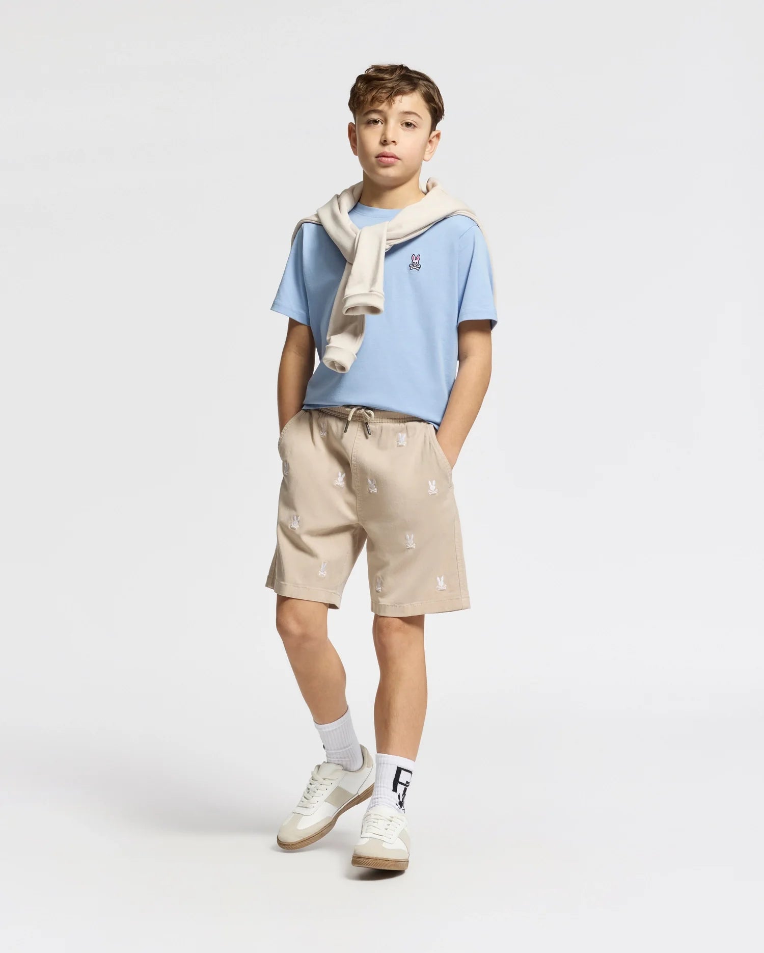 A boy exudes effortless style against a plain backdrop in a light blue t-shirt, white sneakers, and the Psycho Bunny Kids All Over Willis Stretch Tencel Short in beige with subtle patterns. A beige sweater drapes over his shoulders as he casually rests his hands in his pockets.