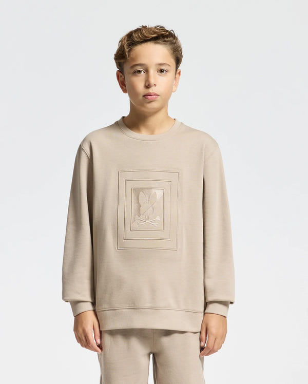 A young person dressed in a beige KIDS OAK CITY CREW NECK SWEATSHIRT by Psycho Bunny, along with matching pants, stands against a plain white background. This sweatshirt features an embroidered design in the center, combining style and comfort for any casual occasion.