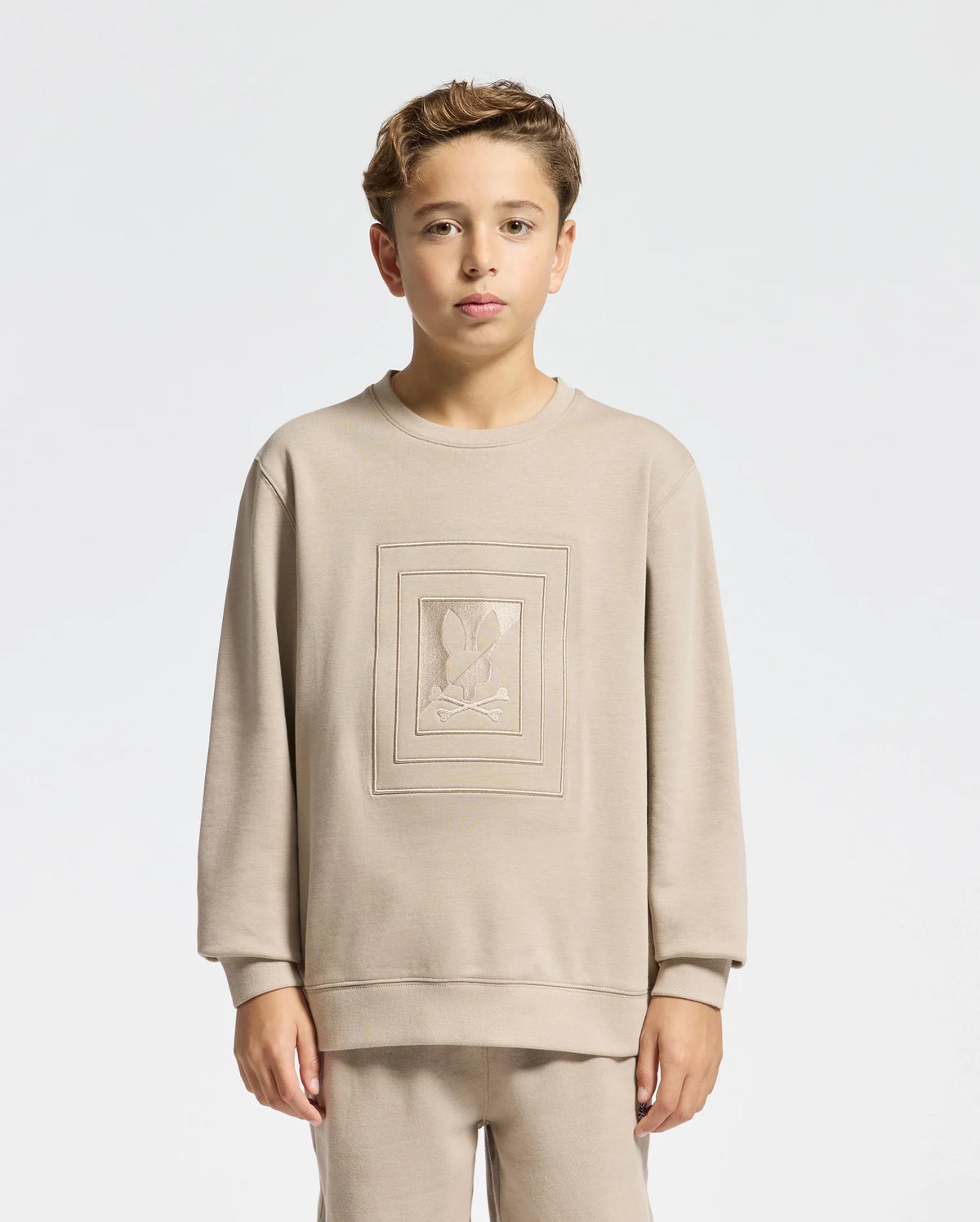 A young person dressed in a beige KIDS OAK CITY CREW NECK SWEATSHIRT by Psycho Bunny, along with matching pants, stands against a plain white background. This sweatshirt features an embroidered design in the center, combining style and comfort for any casual occasion.