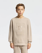 A young person dressed in a beige KIDS OAK CITY CREW NECK SWEATSHIRT by Psycho Bunny, along with matching pants, stands against a plain white background. This sweatshirt features an embroidered design in the center, combining style and comfort for any casual occasion.