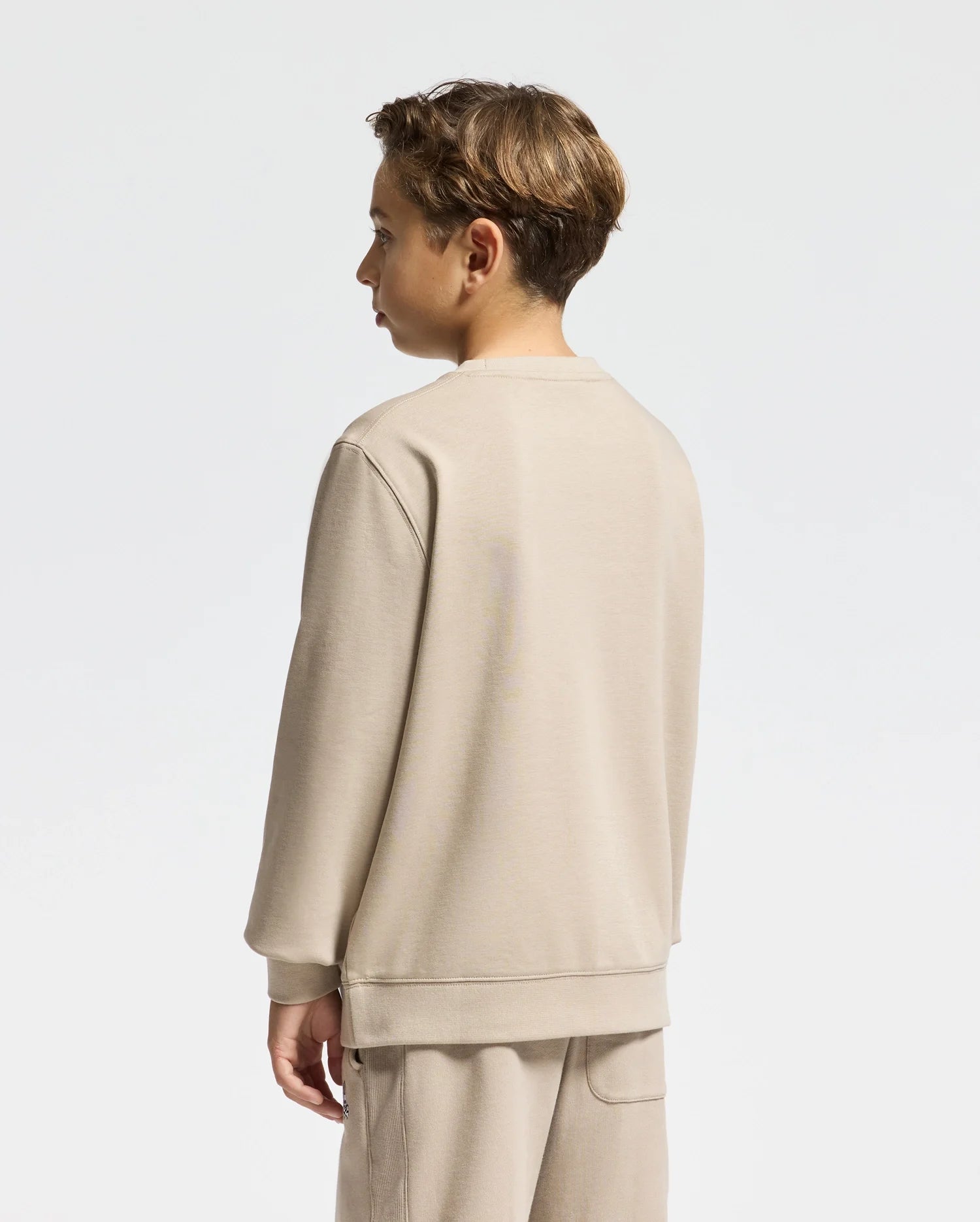 A young person with short brown hair stands facing sideways, wearing a beige KIDS OAK CITY CREW NECK SWEATSHIRT by Psycho Bunny and matching pants. The lightweight outfit contrasts against the plain light gray background.