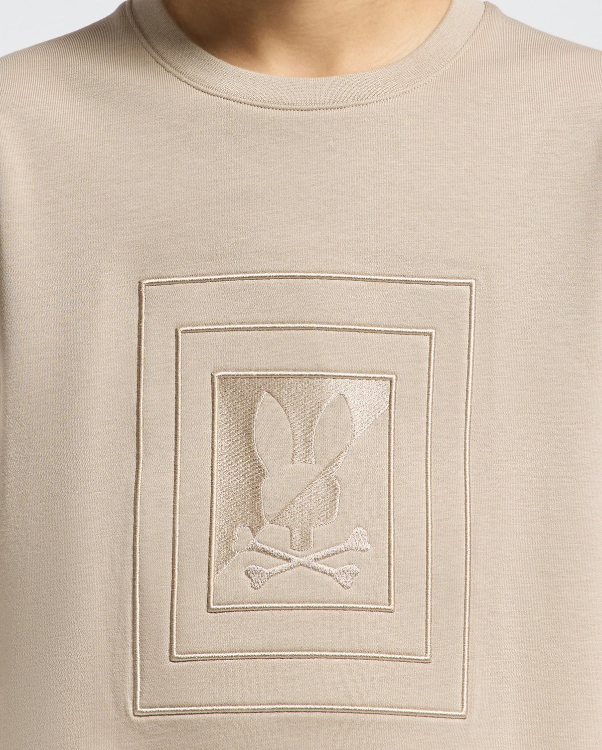 A close-up of the Kids Oak City Crew Neck Sweatshirt in beige, designed by Psycho Bunny, showcases a textured rectangular design. Within this design is an embroidered abstract emblem featuring a rabbit head and crossbones motif, presented in a slightly darker shade of beige.