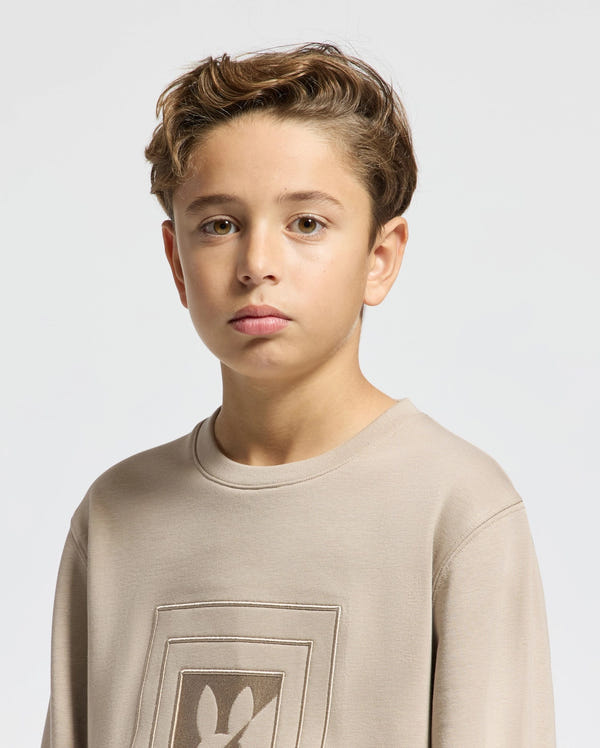 A young boy with brown hair is wearing a beige KIDS OAK CITY CREW NECK SWEATSHIRT by Psycho Bunny. He has a neutral expression and is standing against a plain light background.