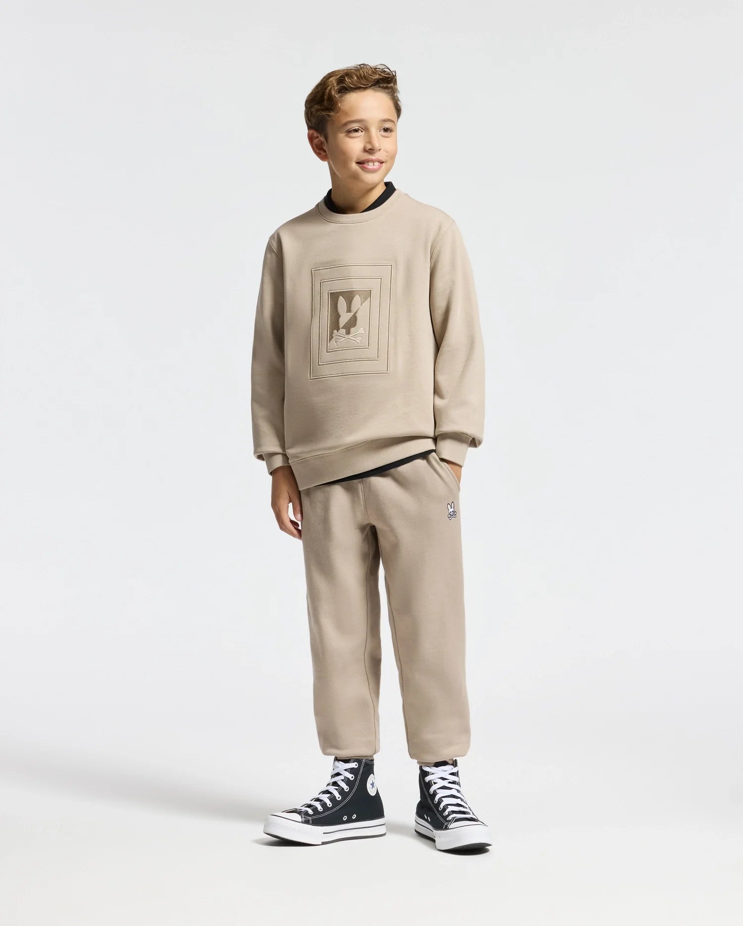A young boy stands smiling, dressed in a beige Psycho Bunny Kids Oak City Crew Neck Sweatshirt and matching pants featuring a design on the chest. He is wearing black high-top sneakers with white soles against a plain white background.