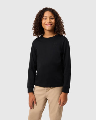 A person with curly hair smiles wearing a Psycho Bunny Kids Alberton long sleeve tee in black, paired with beige pants. Made from Pima cotton, it features a subtle chest design. They stand against a plain light gray background.
