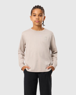 A young person with braided hair wears the Psycho Bunny KIDS ALBERTON LONG SLEEVE TEE (B0T867D200) in light beige, adorned with a rabbit outline on the chest. They rest their hands in black pants pockets, set against a light gray backdrop.