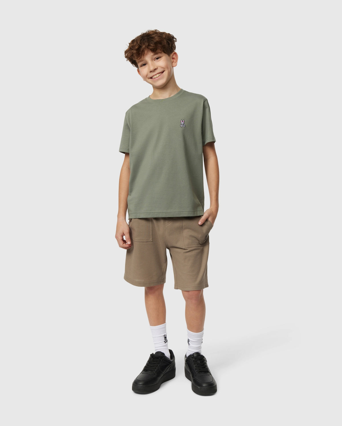 A curly-haired boy smiles in a Psycho Bunny KIDS CLASSIC CREW NECK TEE, brown shorts, white socks, and black sneakers. He casually stands with one hand in his pocket against a plain light background, ready for his back-to-school adventure.