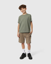 A curly-haired boy smiles in a Psycho Bunny KIDS CLASSIC CREW NECK TEE, brown shorts, white socks, and black sneakers. He casually stands with one hand in his pocket against a plain light background, ready for his back-to-school adventure.