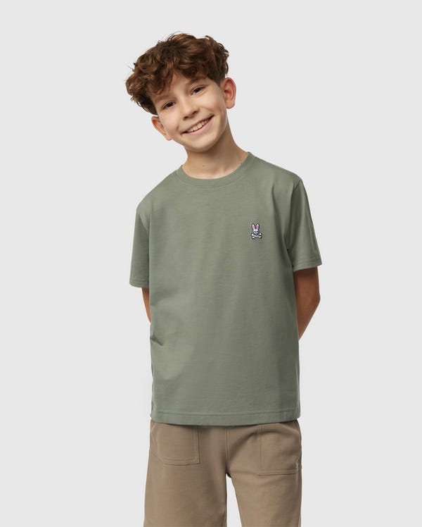 A smiling boy with curly hair stands hands behind his back, wearing a Psycho Bunny kids' classic crew neck tee in green (B0U014B200), made of 100% Pima cotton, and beige pants. The backdrop is neutral light grey—an ideal back-to-school outfit.