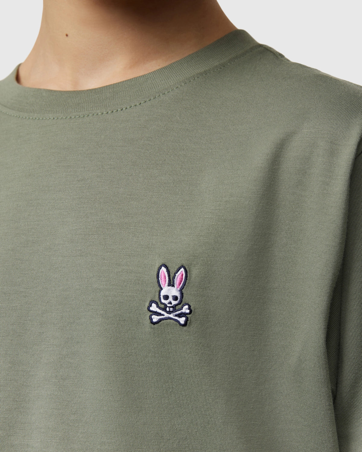 Close-up of a kid wearing the Psycho Bunny Kids Classic Crew Neck Tee in green. Perfect for back-to-school, this 100% Pima cotton tee features an embroidered bunny-headed skull and crossbones logo on the chest.